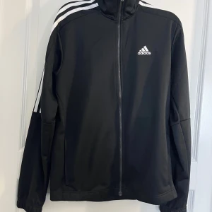Svart träningsjacka från Adidas - Säljer en svart Adidas träningsjacka med vita ränder längs ärmarna och dragkedja framtill. Jackan har hög krage och klassisk Adidas-logga på bröstet. Perfekt för sportiga outfits eller till vardags.
