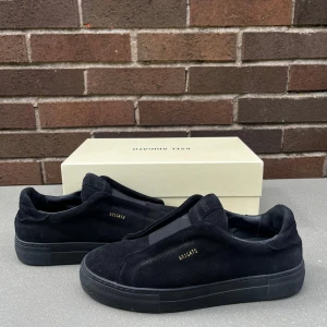 Svarta sneakers från Axel Arigato - Säljer ett par svarta sneakers från Axel Arigato i mocka med diskret logga på sidan. Skorna har en platt sula och rund tå, samt minimalistisk design utan snörning. Perfekta för en stilren look.