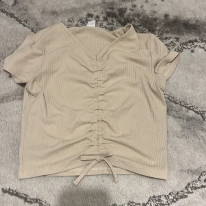 Beige ribbad topp med knyt från Lindex - Säljer en beige ribbad topp från Lindex med v-ringning och knyt framtill. Toppen har korta ärmar och en snygg rynkdetalj längs mitten. Perfekt för en stilren och avslappnad look.