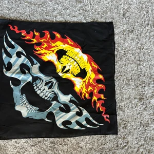 Svart halsduk med dödskalle och eld-motiv 50 x 50 cm - Cool svart halsduk med stort tryck av en dödskalle i grått och vitt, omgiven av eldflammor i rött, gult och orange. Perfekt för dig som gillar tuffa accessoarer och vill sticka ut. Bomull. Mått 50 x 50 cm. Helt ny.