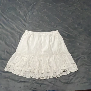 Vit kjol med broderade detaljer - Söt vit kjol med bred volangkant och broderade hålmönster. Kjolen har resår i midjan och är perfekt för varma dagar. Den är kort och har en romantisk känsla tack vare de fina broderierna.