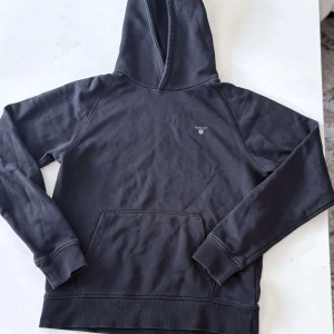 Svart hoodie från Gant - Svart hoodie från Gant med liten logga på bröstet. Klassisk modell med huva och magficka, ribbade muddar vid ärmslut och nederkant. Perfekt för en avslappnad stil. Använd 2-3 gånger! Pris kan diskuteras!😉