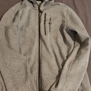 Ljusgrå hoodiejacka från Sail Racing - Ljusgrå hoodiejacka från Sail Racing med dragkedja framtill och en bröstficka med dragkedja. Jackan har huva och logga på bröstet. Perfekt för lager-på-lager och vardagsstil.