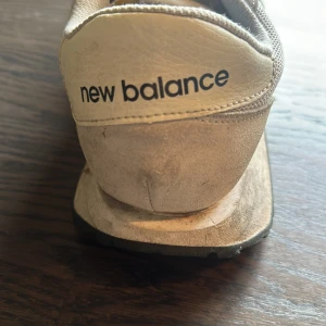 Vita sneakers från New Balance - Säljer ett par vita sneakers från New Balance med svart N-logga på sidan. Skorna har snörning och är tillverkade i syntetmaterial och mesh. Perfekta för dig som gillar klassisk och sportig stil.