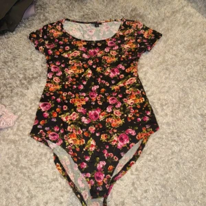 Blommig svart bodysuit från Amisu - Säljer en svart bodysuit från Amisu med färgglatt blommönster i rosa, orange och grönt. Modellen har rund halsringning och korta ärmar. Perfekt för dig som vill ha en färgstark och bekväm topp.