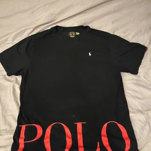 Svart t-shirt från Polo Ralph Lauren - Svart t-shirt från Polo Ralph Lauren med stor röd POLO-text längst ner och liten broderad logga på bröstet. Klassisk rund hals och korta ärmar. Perfekt för en stilren och sportig look. Storlek XL men passar M aldrig använd så helt ny