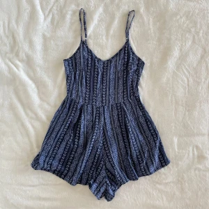 Bluesteel blå mönstrad playsuit med smala axelband - Supersöt blå playsuit med smala axelband och v-ringning. Playsuiten har ett diskret blommigt mönster i vitt och blått, samt en luftig passform som är perfekt för varma dagar.