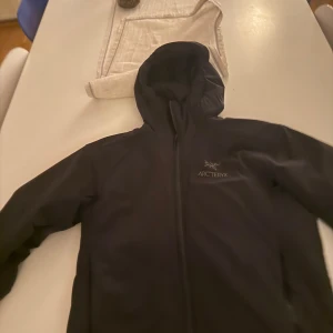 Svart vindjacka från Arc'teryx med huva - Säljer en svart vindjacka från Arc'teryx med huva och dragkedja framtill. Jackan har en stilren design och är perfekt för utomhusaktiviteter. Logotypen syns på bröstet. Passar dig som gillar funktionella och snygga plagg. Modellen heter atom ar och är perfekt till diverse olika aktiviteter. Storlek är S passar även M