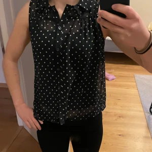 Mörkblå ärmlös blus med vita prickar från Holly & Whyte - Säljer en mörkblå ärmlös blus från Holly & Whyte med vita prickar och liten krage. Blusen är tunn och luftig, perfekt för varma dagar. Stängs med knappar framtill och det sitter fast ett linne under. 💕 