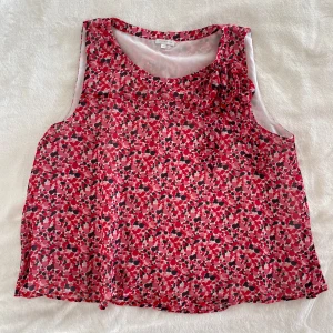 Blommig ärmlös blus med rosett - Säljer en luftig och ärmlös blus från Stockh LM med blommigt mönster i rött, rosa och svart. Blusen har en söt rosettdetalj vid halsen och är tillverkad i ett tunt, syntetiskt material. Perfekt för varma dagar!