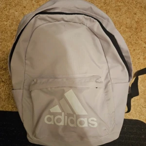Ljusgrå ryggsäck från Adidas - Säljer en ljusgrå ryggsäck från Adidas med stort vitt logotryck på framsidan. Väskan har ett stort huvudfack, en mindre ytterficka med dragkedja och justerbara axelremmar. Perfekt för skola eller träning.