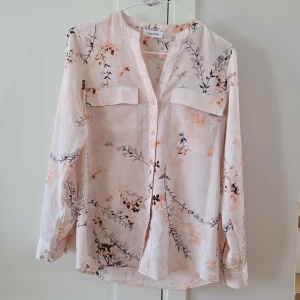 Ljusrosa blommig skjorta från Calvin Klein - Säljer en ljusrosa skjorta från Calvin Klein med blommigt mönster i svart, grått och persikofärgat. Skjortan har lång ärm, två bröstfickor och knappar framtill. 