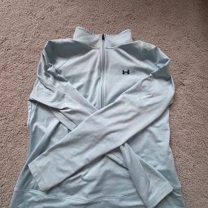 Ljusblå långärmad träningströja från Under Armour - Säljer en ljusblå långärmad träningströja från Under Armour med halv dragkedja framtill och hög krage. Perfekt för löpning eller gym. Tröjan har en stilren design med mörkblå logga på bröstet och ryggen.