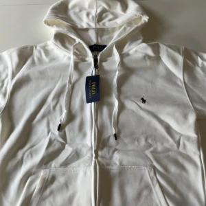 Vit zip hoodie från Polo Ralph Lauren - Vit hoodie med dragkedja från Polo Ralph Lauren. Klassisk design med huva och snörning, samt broderad logga på bröstet. Perfekt för en avslappnad och stilren look. Helt ny,(oanvänd)