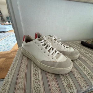 Hugo boss skor - Säljer ett par vita sneakers från BOSS med röd detalj på hälen och mocka vid tån. Skorna har snörning och en platt sula. Perfekta för dig som gillar stilrena och klassiska sneakers med en twist.