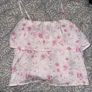 Vit blommig linnetopp med broderade detaljer - Söt vit linnetopp med rosa blommor samt broderade håldetaljer. Toppen har smala axelband och volang upptill, perfekt för varma dagar. Ganska kort, men superfin💕