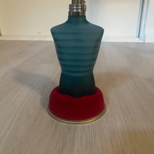 Le Male Eau de Toilette från Jean Paul Gaultier - Ikonisk herrparfym i blå, matt glasflaska formad som en manlig torso med horisontella ränder. Flaskan står på en rund röd sammetsbas och har metallock. Doften är fräsch och maskulin, perfekt för dig som gillar klassiska parfymer.