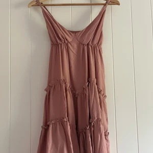 Rosa volangklänning med smala axelband - Säljer en romantisk rosa klänning med smala axelband och flera volanglager. Klänningen har en lös passform och är perfekt för varma dagar. Den har en mjuk, flowy känsla och v-ringad halsringning.