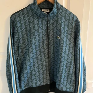 Mönstrad blå track jacket från Lacoste - Säljer en blå track jacket från Lacoste med geometriskt mönster och dragkedja. Jackan har vita och blå ränder längs ärmarna och klassisk Lacoste-logga på bröstet. Perfekt för en sportig och avslappnad stil. Köpt i Frankrike helt ny nästan använd en gång och de var när jag väl köpte den. Har ej sätt någon ha på sig denna eller något vilket gör den unik.