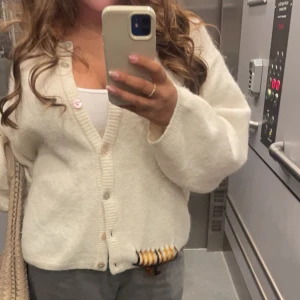 Vit stickad kofta med knappar - Mysig vit stickad kofta med v-ringning och knäppning framtill. Koftan har långa ärmar och en avslappnad passform, perfekt att slänga över en topp. Klassisk och enkel design som passar till mycket.