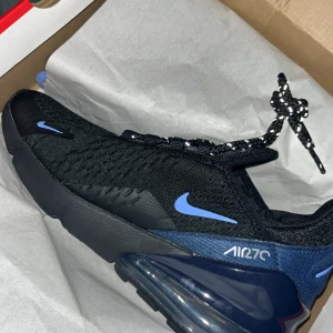 Nike Air Max 270 sneakers svart/blå - Säljer ett par Nike Air Max 270 sneakers i svart med blå detaljer och sula. Skorna har en luftig ovandel i syntetmaterial, snörning och en tydlig Air Max-dämpning i hälen. Perfekta för dig som vill ha både stil och komfort. Endast använda en gång! Passar en storlek större! Skicka bud!