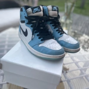 Nike Air Jordan 1 Retro High OG University Blue - Nike Air Jordan 1 Retro High OG i färgen University Blue med svart swoosh och vita paneler. Skorna har högt skaft, svarta skosnören och klassisk Air Jordan-logga på sidan. Ovandelen är i skinn och mocka för en snygg kontrast.