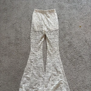 Vita utsvängda spetsbyxor - Säljer ett par vita utsvängda byxor i genombruten spets med blommigt mönster. Byxorna har hög midja och är stretchiga med en riktigt snygg 70-talsvibe. Perfekta för dig som vill sticka ut lite extra! dock ett litet hål mellan benen men inget man ser