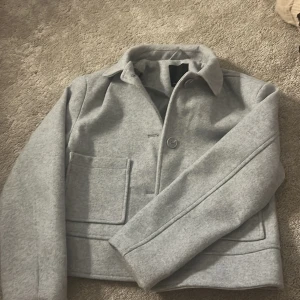 Ljusgrå kappa från 157 - Säljer en stilren ljusgrå kappa från 157 med klassisk krage och stora framfickor. Jackan har en enkel knäppning framtill och är i ett mjukt ull-liknande material. Perfekt för dig som gillar minimalistisk stil.