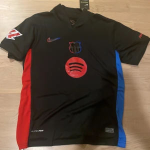 FC Barcelona svart bortatröja Nike Lamine Yamal 19 - Säljer en svart FC Barcelona bortatröja från Nike med Lamine Yamal 19 på ryggen. Tröjan har röda och blå detaljer på sidorna, klubbmärke och sponsorloggor på bröstet. Kortärmad modell i Dri-FIT material.