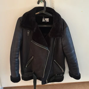 Svart skinnjacka från NA-KD - Snygg svart skinnjacka från NA-KD med mjuk pälsdetalj på kragen. Jackan har en asymmetrisk dragkedja och två fickor med dragkedjor. Perfekt för kyligare dagar med sin stilrena design och bekväma passform.