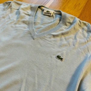 Ljusblå tröja från Lacoste - Säljer en ljusblå tröja från Lacoste med v-ringning och det klassiska krokodilmärket på bröstet.
