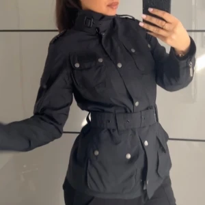 Svart jacka från Barbour - Säljer en stilren svart jacka från Barbour med bälte i midjan och flera fickor. Jackan har en klassisk krage och knappar framtill. Insidan har ett rutigt mönster som ger en snygg kontrast. Perfekt för dig som vill ha en tidlös look.                       Den passar storlek 36 då jag själv har storlek 36 i vanligt fall. 
