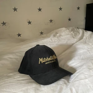 Svart keps från Mitchell & Ness - Snygg svart keps från Mitchell & Ness med guldfärgad broderad text på framsidan. Justerbar passform med snapback-stängning. Perfekt för en casual look.