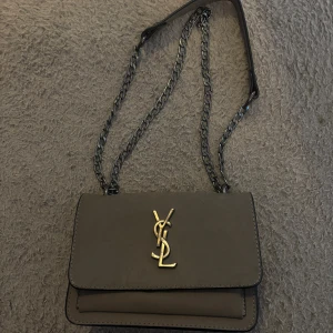 Grå axelväska från Yves Saint Laurent - Snygg svart axelväska från Yves Saint Laurent med kedjerem och ikonisk logotyp på framsidan. Väskan har en stilren design och är perfekt för att bära dina nödvändigheter. Insidan har en praktisk dragkedja för extra säkerhet.