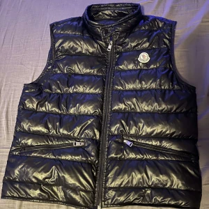 Svart dunväst från Moncler - Snygg svart dunväst från Moncler med dragkedja och två praktiska fickor framtill. Västen har en stilren design med Moncler-logga på bröstet. Perfekt för kyligare dagar när du vill hålla stilen. Man får även med en moncler påse, sjävklart äkta. Priset går att diskutera 