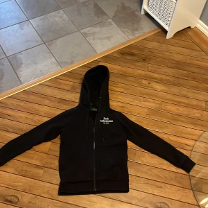 Svart hoodie från Peak Performance - Säljer en stilren svart hoodie från Peak Performance med dragkedja och broderad logga på bröstet. Perfekt för en avslappnad look. Den har långa ärmar och en praktisk huva.