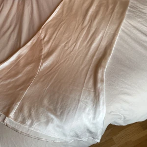 Vit långklänning från Gina Tricot - Elegant vit långkjok från Gina Tricot i satin. Kjolen har en enkel och stilren design, perfekt för speciella tillfällen. Den är axelbandslös och har en lätt utsvängd form som ger en vacker siluett. Den är i storlek M