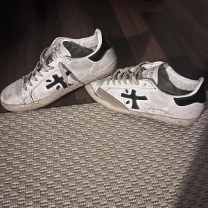 Unika vita sneakers från Premiata - Snygga vita sneakers från Premiata med svarta detaljer och snörning. Perfekta för en stilren look. Skorna har en klassisk design med en bekväm passform. Skolan är i begnagnat skick och respektive häl är skadad (se bild 5). Box medföljer!