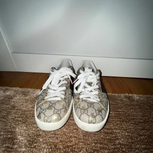 Gucci ace sneakers med monogram - Snygga Gucci sneakers med det klassiska monogrammönstret. Skorna har vita snören och en vit sula. De har gröna och röda ränder på sidan samt metallic-detaljer i rött och grönt på hälen. Perfekta för att ge din outfit en lyxig touch. Skorna har en liten defekt som man kan se på 3de och 5de bilden och därför jag säljer för lite lägre pris. Om man bortser från de så finns de inga defekter. Skorna ligger på 7-8k nypris och jag säljer för 1549kr😊😊