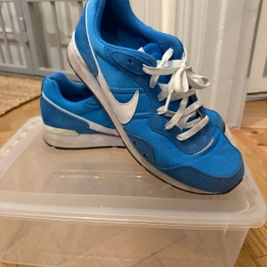 Blå Nike sneakers - Säljer ett par blå Nike sneakers med vita detaljer och snörning. Skorna har en klassisk design med en vit Nike-logga på sidan och är perfekta för en sportig look. Storlek 45