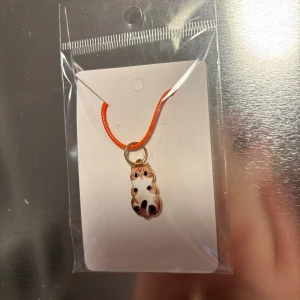 Halsband med kattberlock - Charmigt halsband med en söt kattberlock i olika färger. Berlocken hänger på ett orange band och har detaljer i guld. Perfekt för kattälskare som vill ha en unik accessoar.