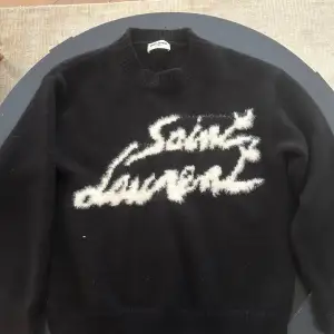 Säljer en svart tröja från Saint Laurent med vit text på framsidan. Tröjan har en rund halsringning och är tillverkad i ett mjukt material. Perfekt för en stilren look. Köpt för ett bra tag sen i Italien! Bara höra av sig vid funderingar!