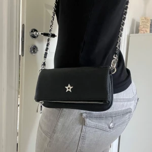Svart axelväska med stjärndetalj från Gina - Snygg svart axelväska från Gina Tricot i skinn med en kedjerem och en glittrig stjärndetalj på framsidan. Väskan har en dragkedja för säker förvaring av dina saker. Perfekt för att ge en stilren touch till din outfit. 