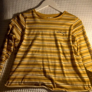 Gul och vit randig långärmad tröja - Säljer en gul och vit randig långärmad tröja med rund hals. Tröjan har ett litet broderat motiv på bröstet. Perfekt för en avslappnad stil.