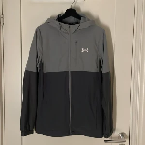 Grå vindjacka från Under Armour - Snygg grå vindjacka från Under Armour med dragkedja och huva. Perfekt för träning eller en aktiv vardag. Jackan har en stilren design med en vit logotyp på bröstet.
