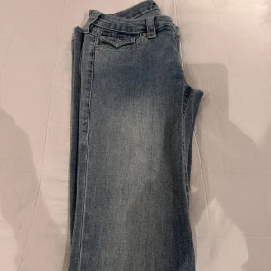 Blå jeans från True Religion - Säljer ett par snygga blå jeans från True Religion med låg midja och flare-stil. De har dekorativa sömmar och knappar på bakfickorna. Perfekta för en avslappnad look.