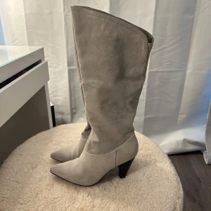 Gråa stövlar med klack - Gråa stövlar i mocka med spetsig tå och låg klack. De har en praktisk dragkedja på sidan för enkel på- och avtagning. Använda endast 1 gång. Storlek 38, passar även 39. Skaftet kan va tajt för dom med breda vader! 