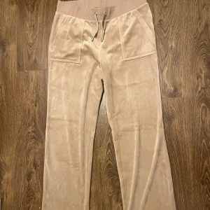 Beige mjukisbyxor från Juicy Couture - Snygga beige mjukisbyxor från Juicy Couture med bred resår i midjan och snörning. Byxorna har fickor både fram och bak med knappdetaljer. Perfekta för en avslappnad stil.