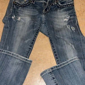 Blå jeans från Miss Me - Snygga blå jeans från Miss Me med slitna detaljer och dekorativa sömmar. Byxorna har en bootcut-stil och är prydda med knappar och broderade bakfickor.