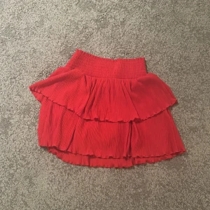 Röd volangkjol från Kappahl - Säljer en söt röd volangkjol från Kappahl. Kjolen har en elastisk midja och två lager volanger som ger en lekfull look. Perfekt för att liva upp garderoben med lite färg!